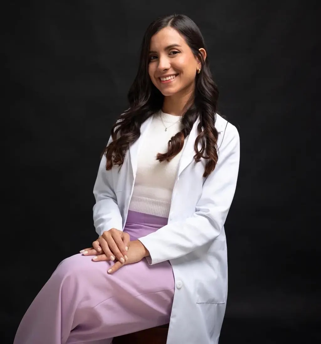 La Dra. Paola Ruiz con bata blanca, sonriendo, sentada frente a un fondo oscuro